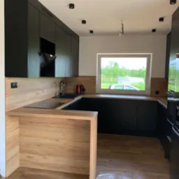 Nowoczesna kuchnia z czarnymi szafkami i drewnianym blatem, widok na ogród przez okno, oświetlenie punktowe na suficie.