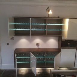 Certa Bartłomiej Bojarski - Montaż mebli kuchennych w trakcie realizacji: szafki z podświetlanymi półkami, widoczne narzędzia i niedokończone wykończenie podłogi.