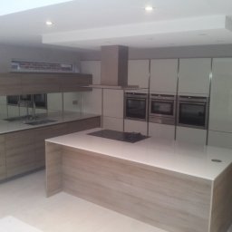 Certa Bartłomiej Bojarski - Nowoczesna kuchnia z wyspą, zabudową meblową w kolorze jasnego drewna i białymi frontami szafek, oświetleniem punktowym i nietypowym oknem nad blatem.