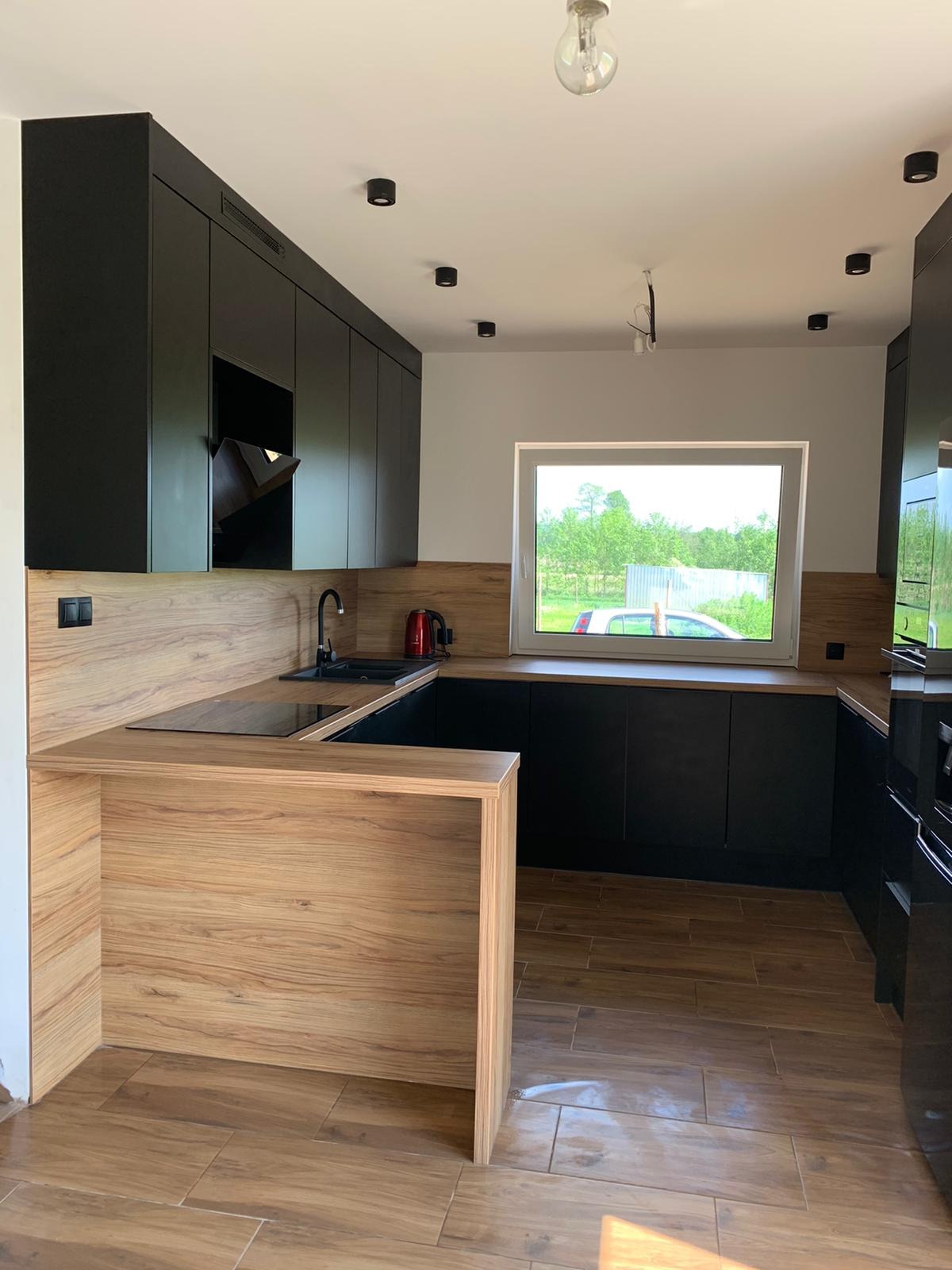 Nowoczesna kuchnia z czarnymi szafkami i drewnianym blatem, widok na ogród przez okno, oświetlenie punktowe na suficie.