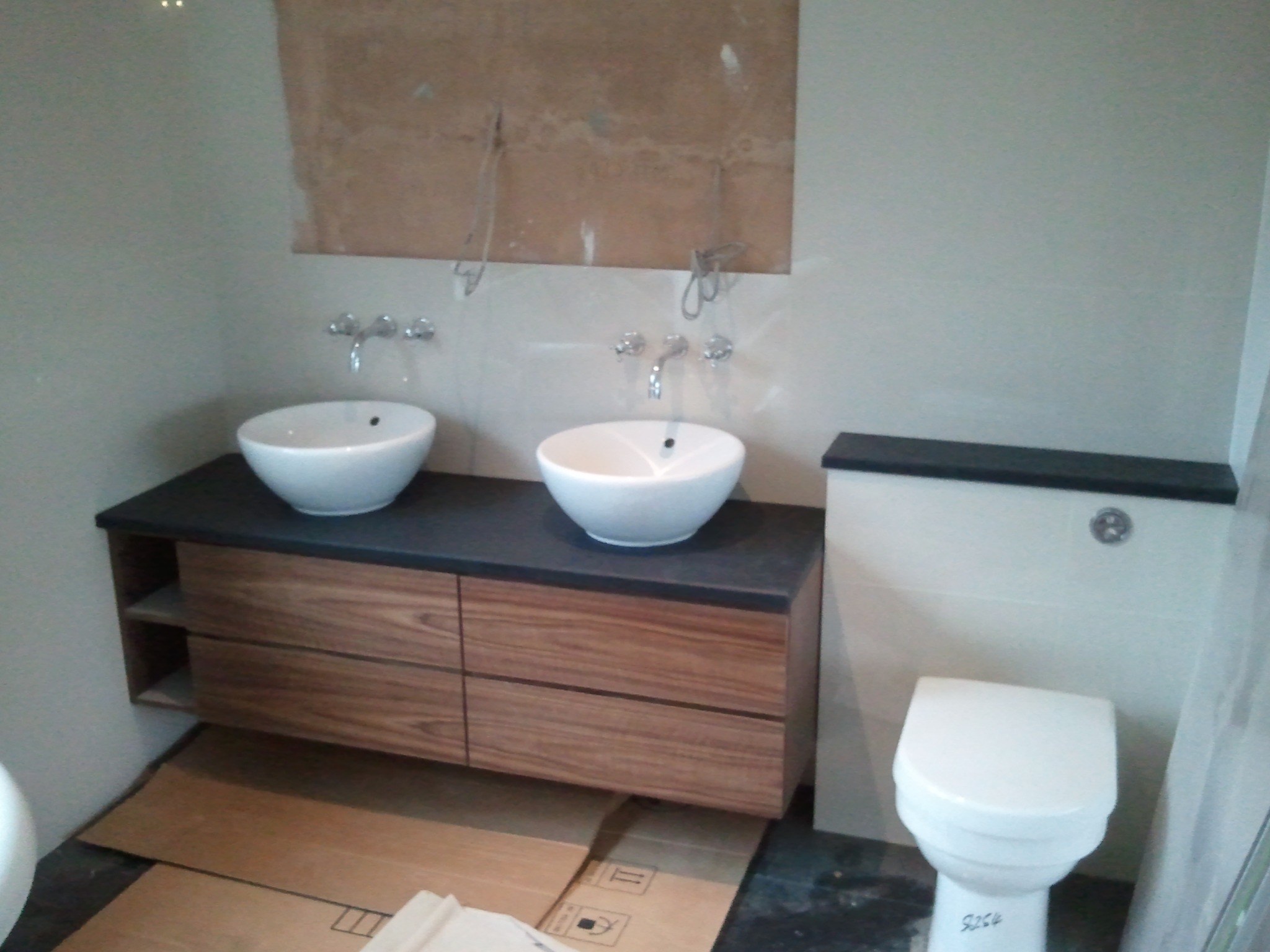 Łazienka w trakcie wykańczania: dwie białe umywalki na drewnianej szafce z czarnym blatem, widoczne kartony ochronne na podłodze, instalacja wodna bez podłączenia armatury, toaleta z czarnym...