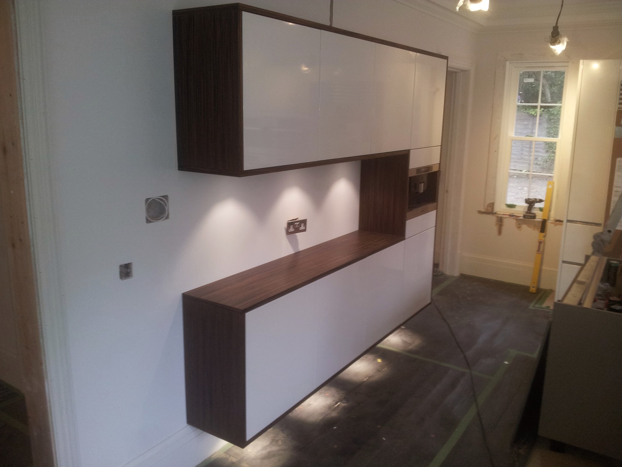 Nowoczesna zabudowa kuchenna z białymi frontami i ciemnobrązowym obramowaniem, oświetlenie punktowe pod szafkami, widoczne gniazdka elektryczne i fragment okna z narzędziami na parapecie.