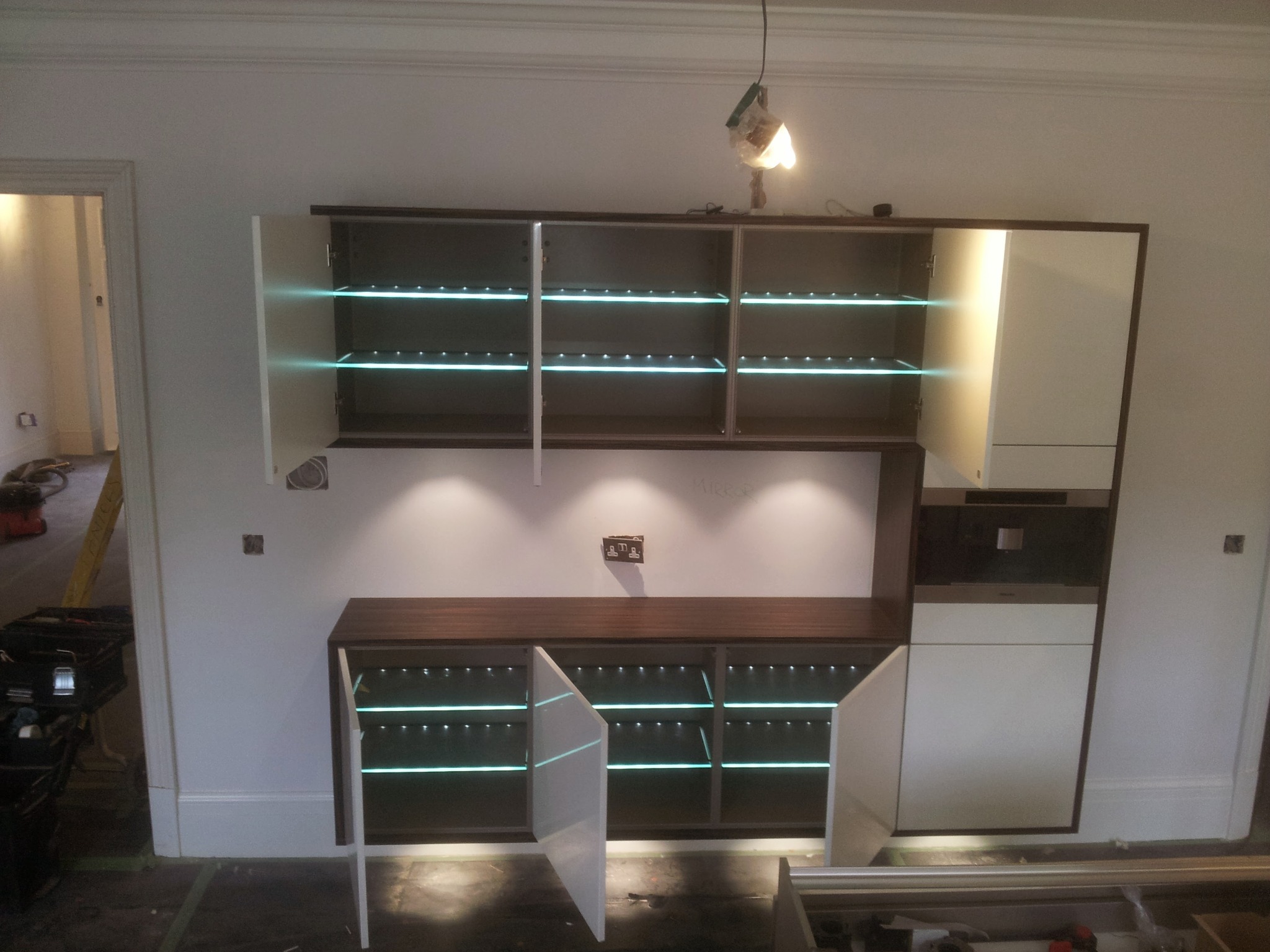 Montaż mebli kuchennych w trakcie realizacji: szafki z podświetlanymi półkami, widoczne narzędzia i niedokończone wykończenie podłogi.