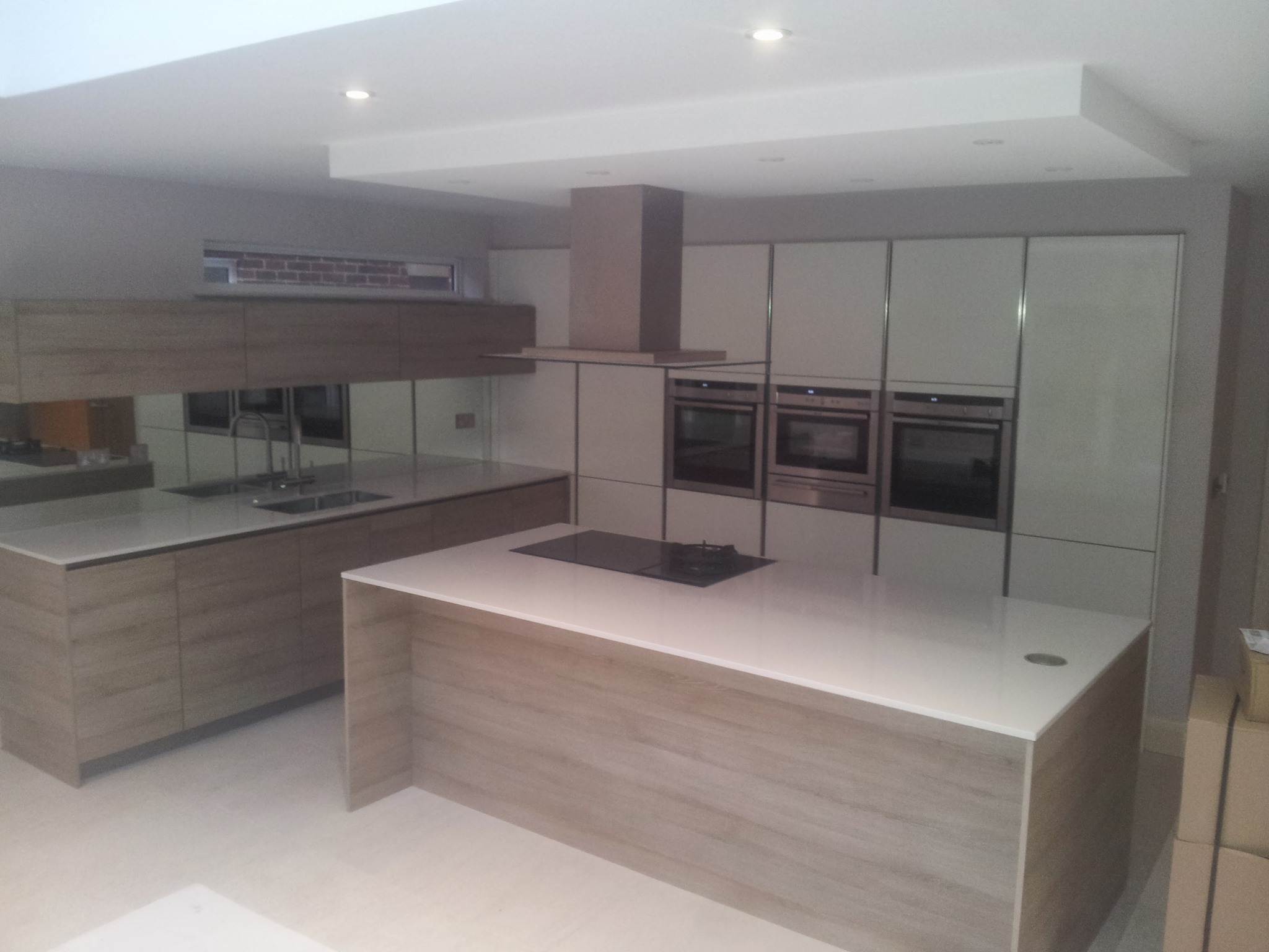 Nowoczesna kuchnia z wyspą, zabudową meblową w kolorze jasnego drewna i białymi frontami szafek, oświetleniem punktowym i nietypowym oknem nad blatem.