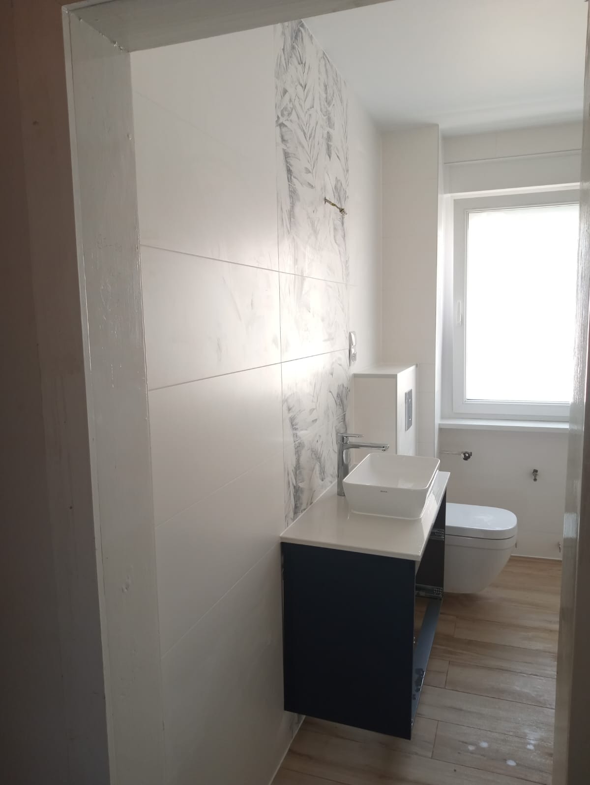 Wykończona łazienka z białymi płytkami imitującymi marmur z szarym wzorem roślinnym, umywalką nablatową na ciemnogranatowej szafce i drewnopodobną podłogą, widok zza futryny.