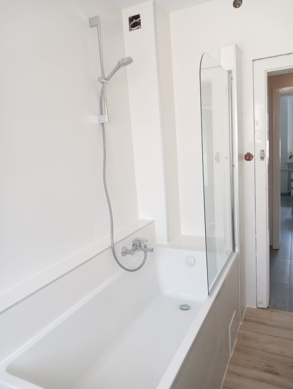 Biała, nowocześnie wykończona łazienka z wanną, szklanym parawanem nawannowym i chromowaną baterią oraz słuchawką prysznicową; widoczne niedokończone prace elektryczne w ścianie.