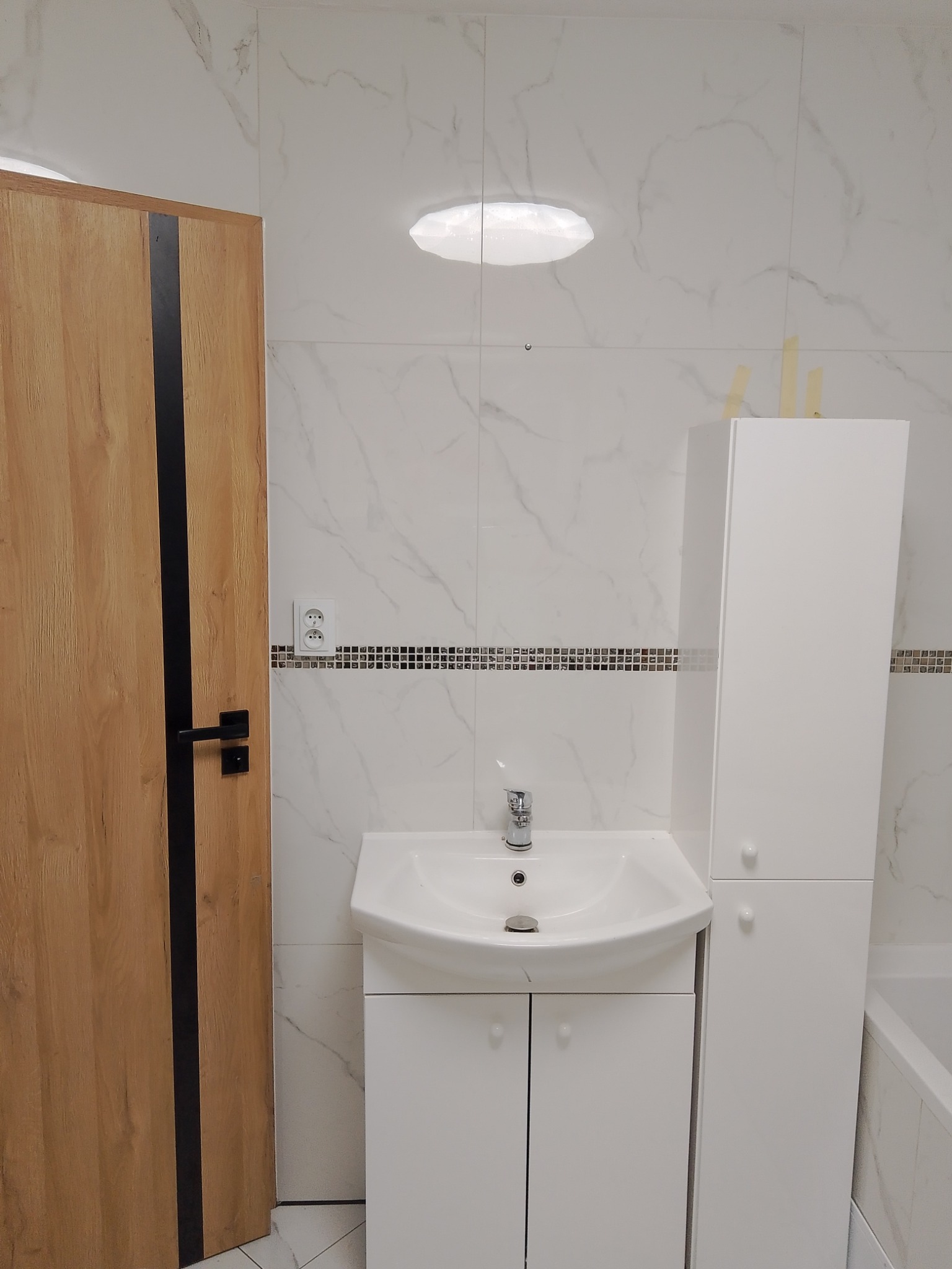 Nowoczesna łazienka z umywalką w szafce, wysoką białą szafką, drzwiami z drewnopodobnym fornirem i czarnym pasem, oraz marmurkowymi płytkami na ścianach.