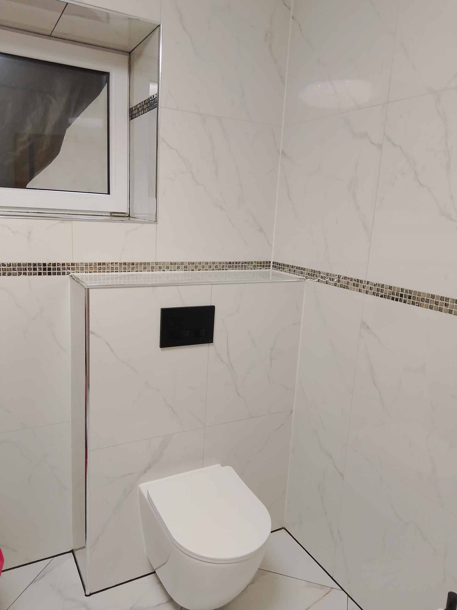 Nowoczesna łazienka z podwieszaną miską WC, białe płytki imitujące marmur z dekoracyjnym paskiem mozaiki, czarny przycisk spłuczki i okno z ramą wykończoną chromem.