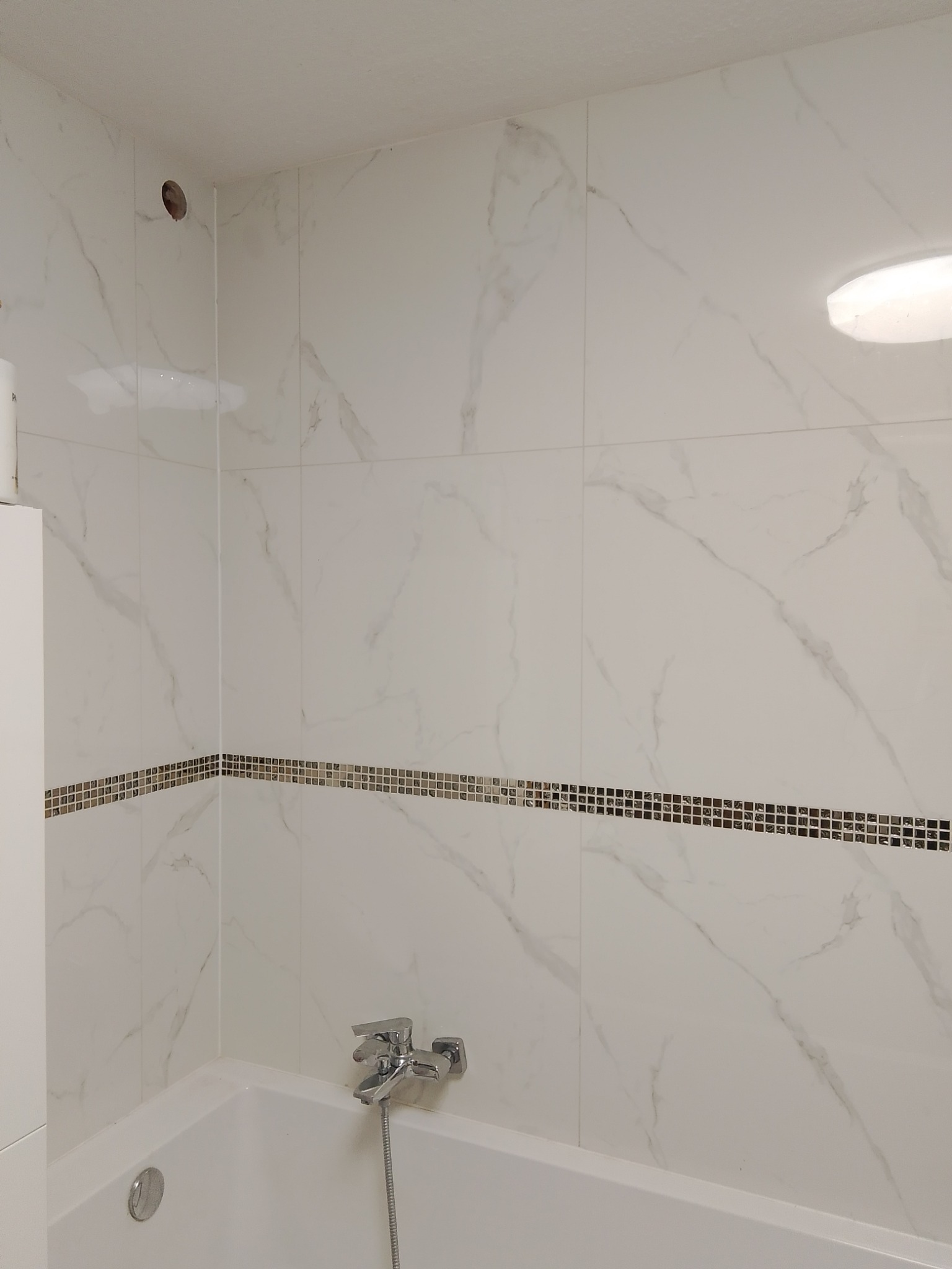 Biała łazienka z wanną, marmurowymi płytkami i dekoracyjnym paskiem mozaiki, widoczne bateria wannowa i otwór w ścianie