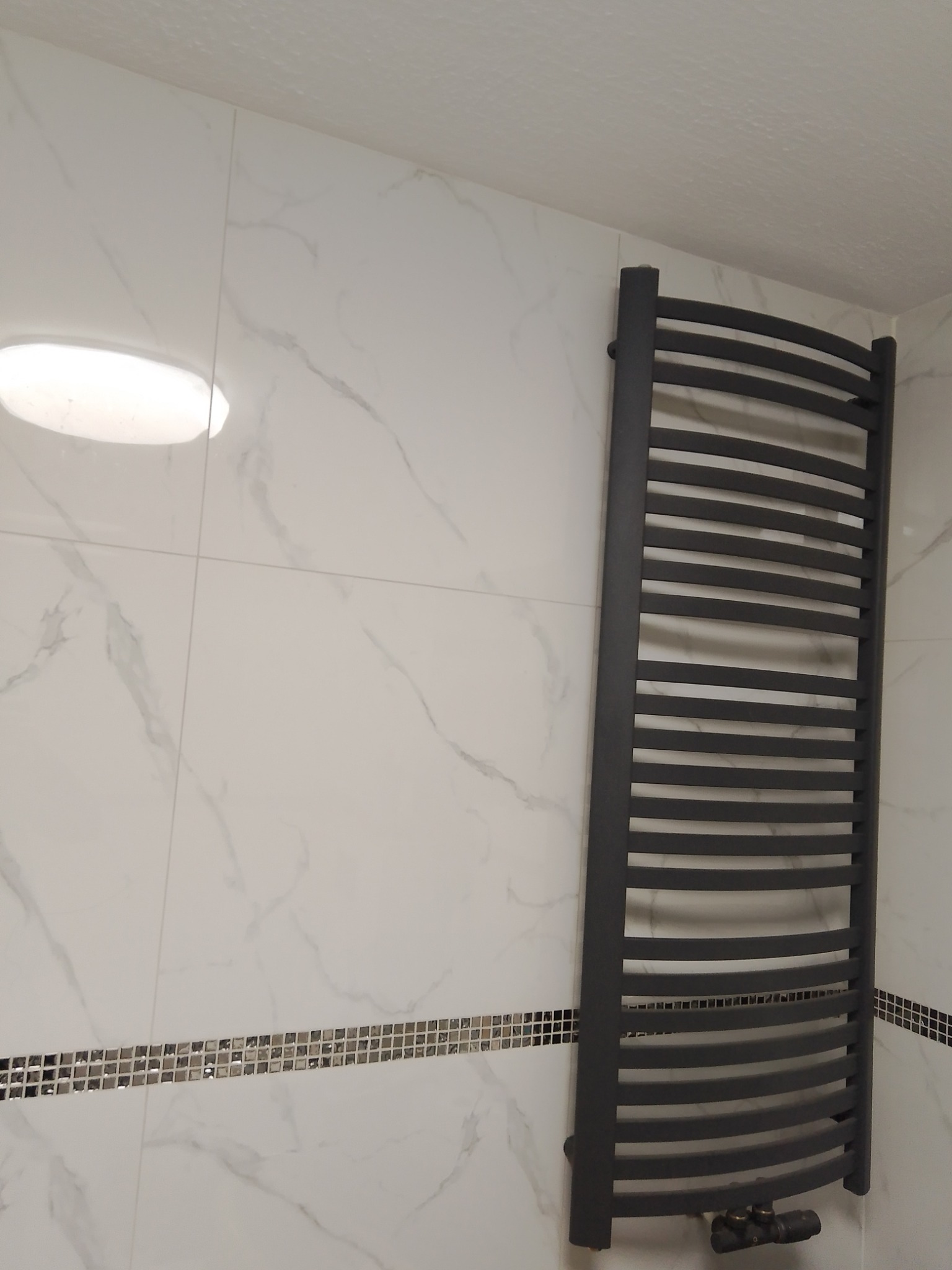 Szary, drabinkowy grzejnik łazienkowy na białej ścianie z marmurowym wzorem i poziomym pasem mozaiki.