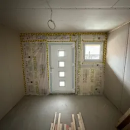 Wnętrze w trakcie prac wykończeniowych: ściany z płyt kartonowo-gipsowych, zamontowane okno i drzwi wejściowe zabezpieczone folią paroizolacyjną z taśmą uszczelniającą, na podłodze leżą drewniane...