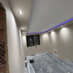 Pokój w trakcie remontu: ściany po szpachlowaniu i gruntowaniu, widoczny fragment surowej cegły, zabezpieczona folią podłoga, zamontowane oświetlenie LED i halogenowe.