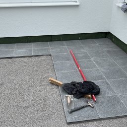 AB Construction - Układanie szarych płytek na zewnątrz budynku, widoczny fragment podłoża przygotowanego pod płytki, poziomica, kielnia i młotek.