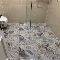 AB Construction - Ukończona łazienka z kabiną prysznicową typu walk-in, wzorzystą podłogą z płytek w odcieniach szarości, beżu i błękitu, beżowymi płytkami ściennymi i częściowo otwartymi białymi drzwiami z ozdobną,...