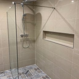 AB Construction - Nowoczesna kabina prysznicowa walk-in z deszczownicą, wykończona beżowymi płytkami imitującymi marmur i dekoracyjną mozaiką podłogową, z wnęką w ścianie.