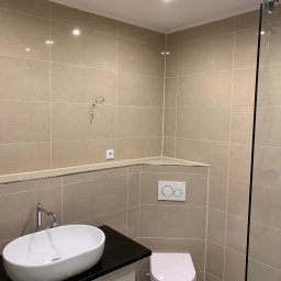 AB Construction - Beżowa łazienka z owalną umywalką nablatową na czarnym blacie, wisząca toaleta i kabina prysznicowa ze szklaną ścianką; widoczne wystające przewody elektryczne.