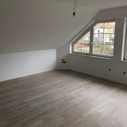 AB Construction - Poddasze po remoncie z jasną podłogą imitującą drewno, świeżo pomalowane ściany na biało, dwa okna z widokiem na jesienne drzewa, widoczne gniazdka elektryczne i wisząca żarówka.