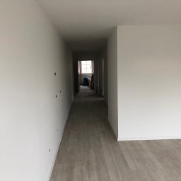 AB Construction - Korytarz w jasnych barwach, świeżo po malowaniu, z widocznymi śladami po otworach na ścianie, podłoga wyłożona panelami imitującymi drewno, w głębi widoczne okno z resztkami materiałów budowlanych.