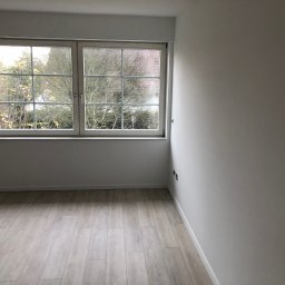 AB Construction - Jasne, nowo wykończone wnętrze pokoju z drewnopodobną podłogą, białymi ścianami i oknem z widokiem na drzewa bez liści.