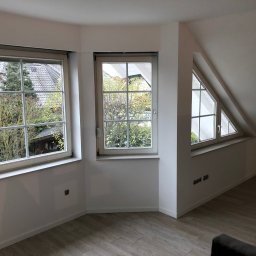 AB Construction - Jasne, wykończone wnętrze poddasza z trzema oknami o różnym kształcie, świeżo pomalowane ściany na biało, panele podłogowe imitujące drewno, widoczne gniazdka elektryczne.