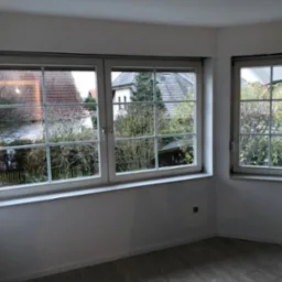 Pusty, świeżo wyremontowany pokój z dwoma oknami z widokiem na domy i drzewa, pomalowany na biało, z panelami podłogowymi o wzorze drewna.