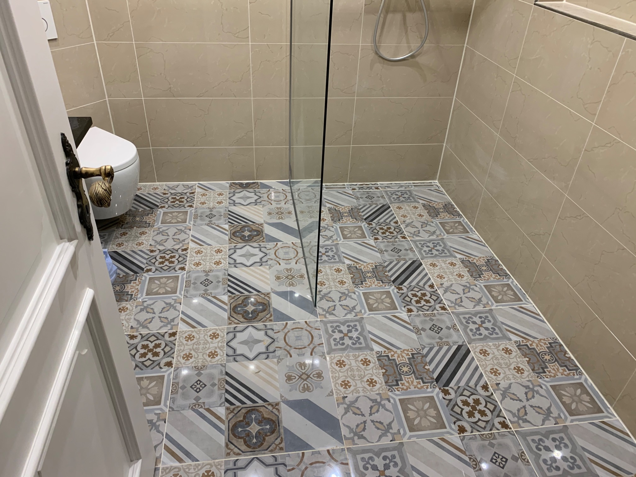 Ukończona łazienka z kabiną prysznicową typu walk-in, wzorzystą podłogą z płytek w odcieniach szarości, beżu i błękitu, beżowymi płytkami ściennymi i częściowo otwartymi białymi drzwiami z ozdobną,...