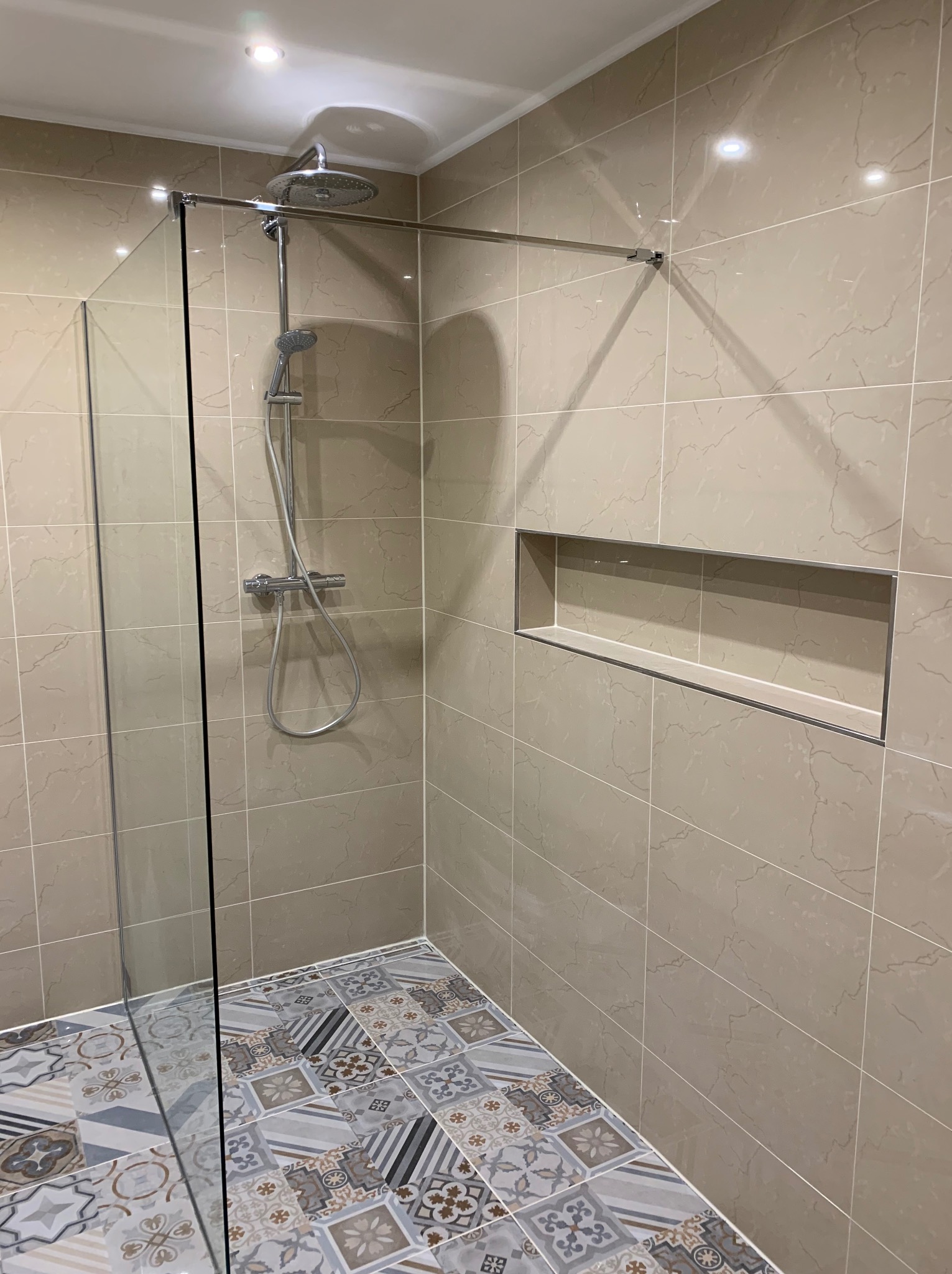 Nowoczesna kabina prysznicowa walk-in z deszczownicą, wykończona beżowymi płytkami imitującymi marmur i dekoracyjną mozaiką podłogową, z wnęką w ścianie.