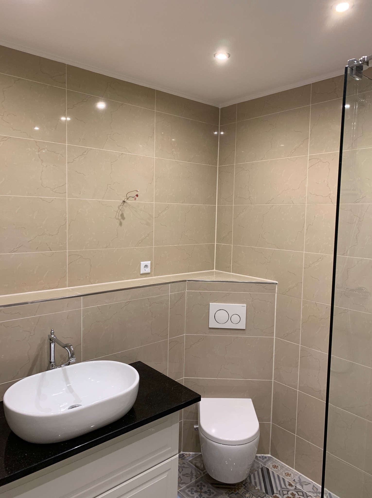 Beżowa łazienka z owalną umywalką nablatową na czarnym blacie, wisząca toaleta i kabina prysznicowa ze szklaną ścianką; widoczne wystające przewody elektryczne.