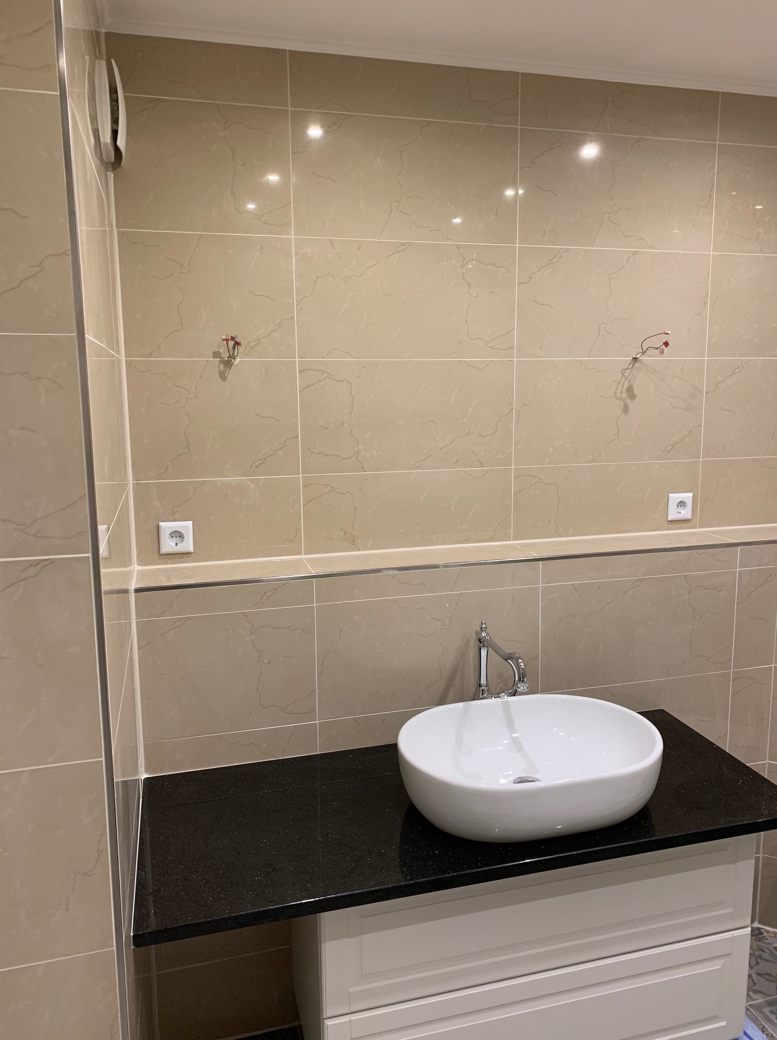 Łazienka w trakcie wykańczania: beżowe płytki, czarny blat z białą umywalką, wystające kable elektryczne, widoczne gniazdko elektryczne.
