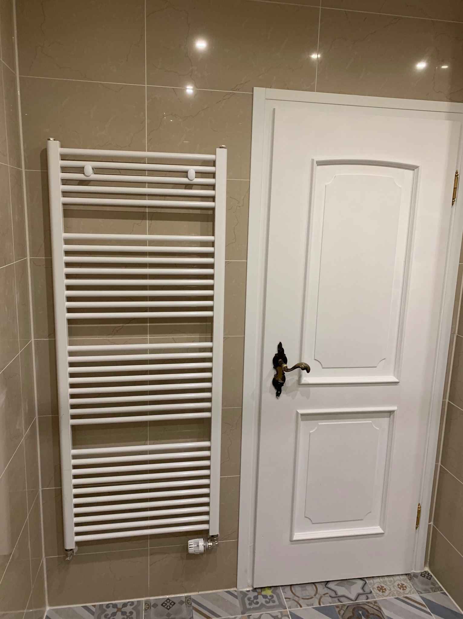 Biały grzejnik drabinkowy zamontowany na beżowej ścianie z płytek, obok białe drzwi z ozdobną, ciemną klamką w wykończonej łazience.