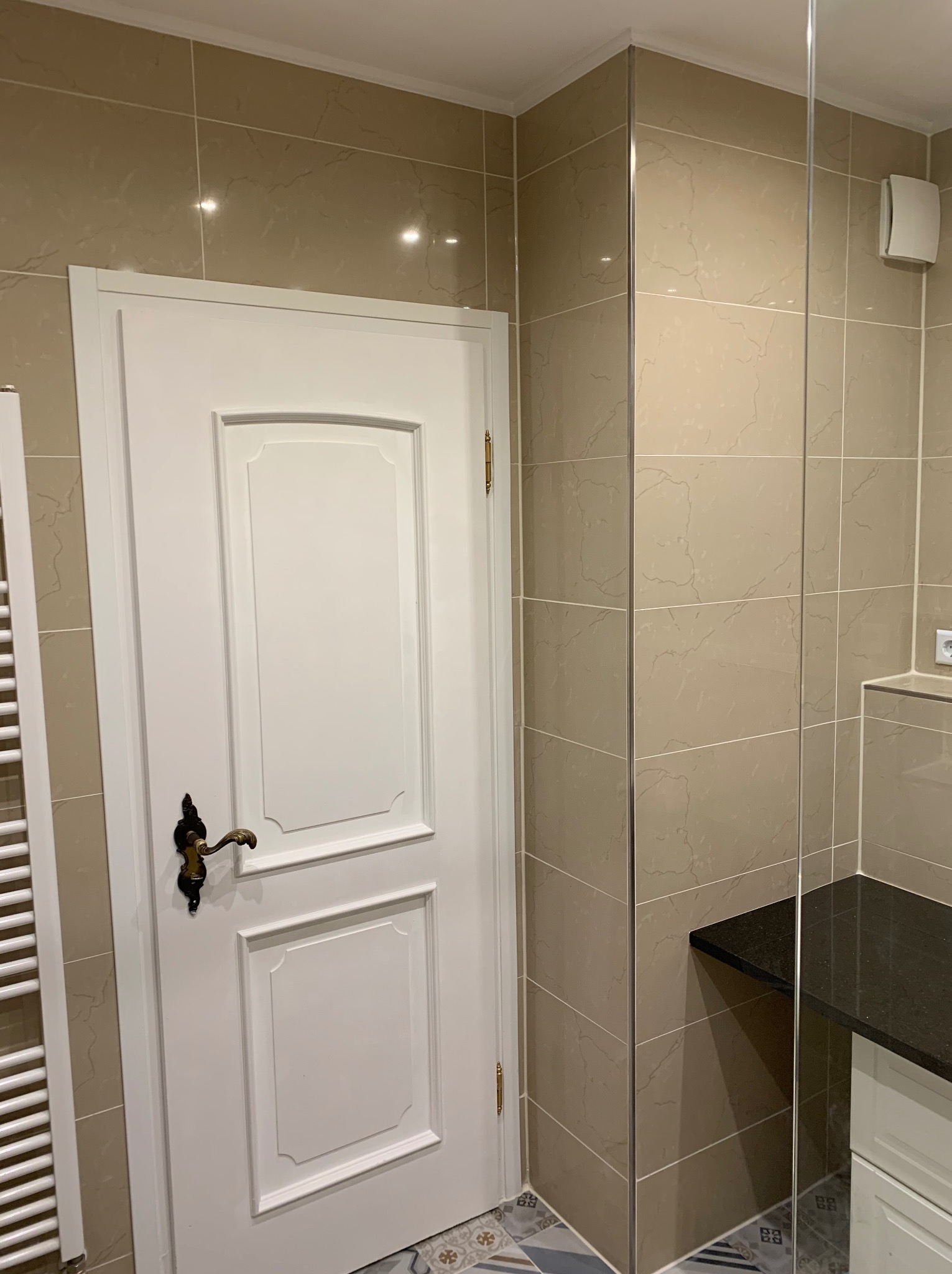 Wykończona łazienka z beżowymi płytkami imitującymi marmur, białe drzwi z ozdobną klamką w stylu retro, grzejnik drabinkowy i fragment blatu z ciemnego kamienia.