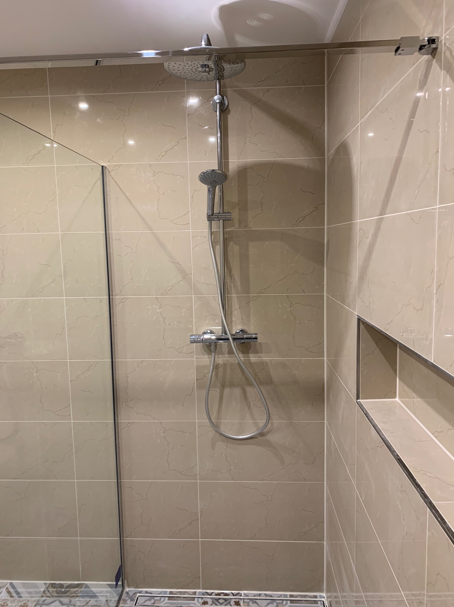 Nowoczesna kabina prysznicowa typu walk-in z deszczownicą i baterią termostatyczną, wykończona beżowymi płytkami imitującymi marmur, z wnęką na kosmetyki.