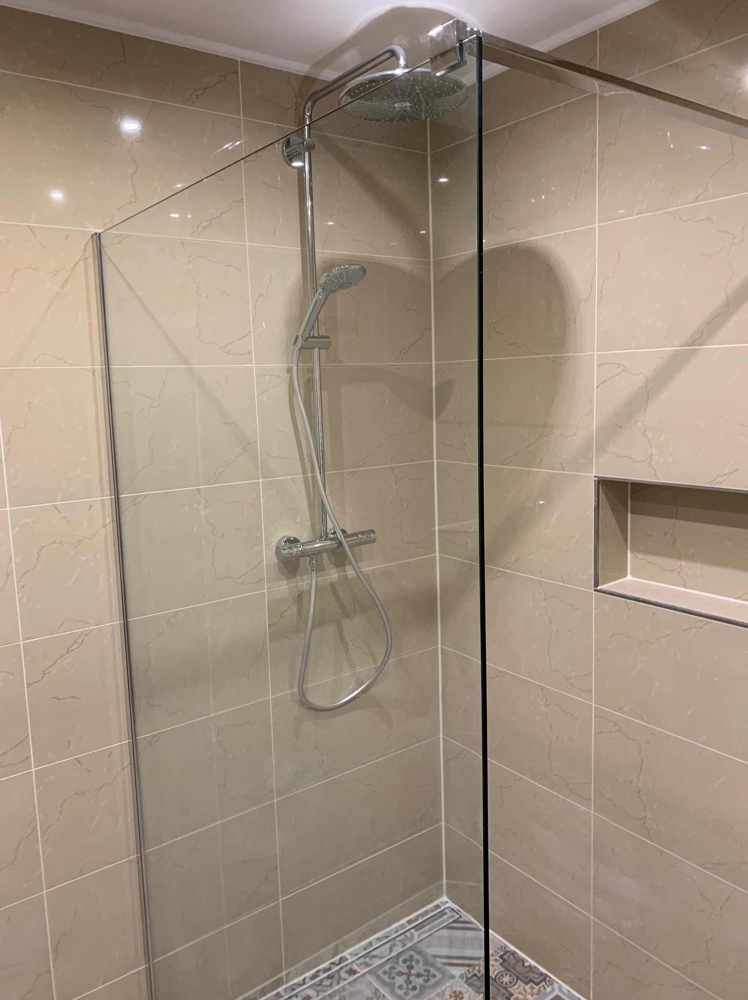 Kabina prysznicowa walk-in ze szklaną ścianką, wykończona beżowymi płytkami imitującymi marmur, z wnęką na kosmetyki i dekoracyjną mozaiką podłogową.