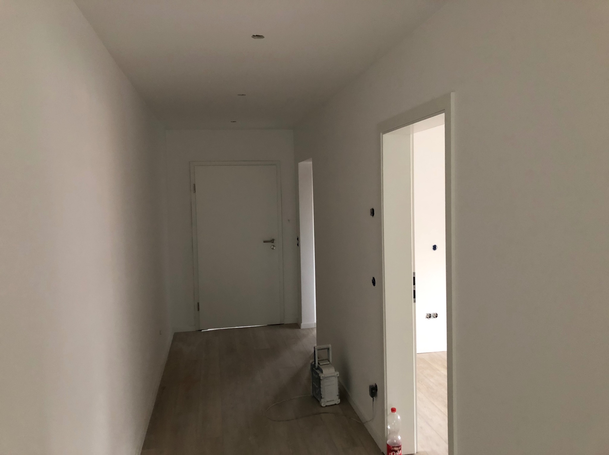 Wnętrze korytarza z białymi ścianami po malowaniu, widoczna podłoga panelowa, białe drzwi, niedokończone gniazdka elektryczne oraz butelka z napojem na podłodze.