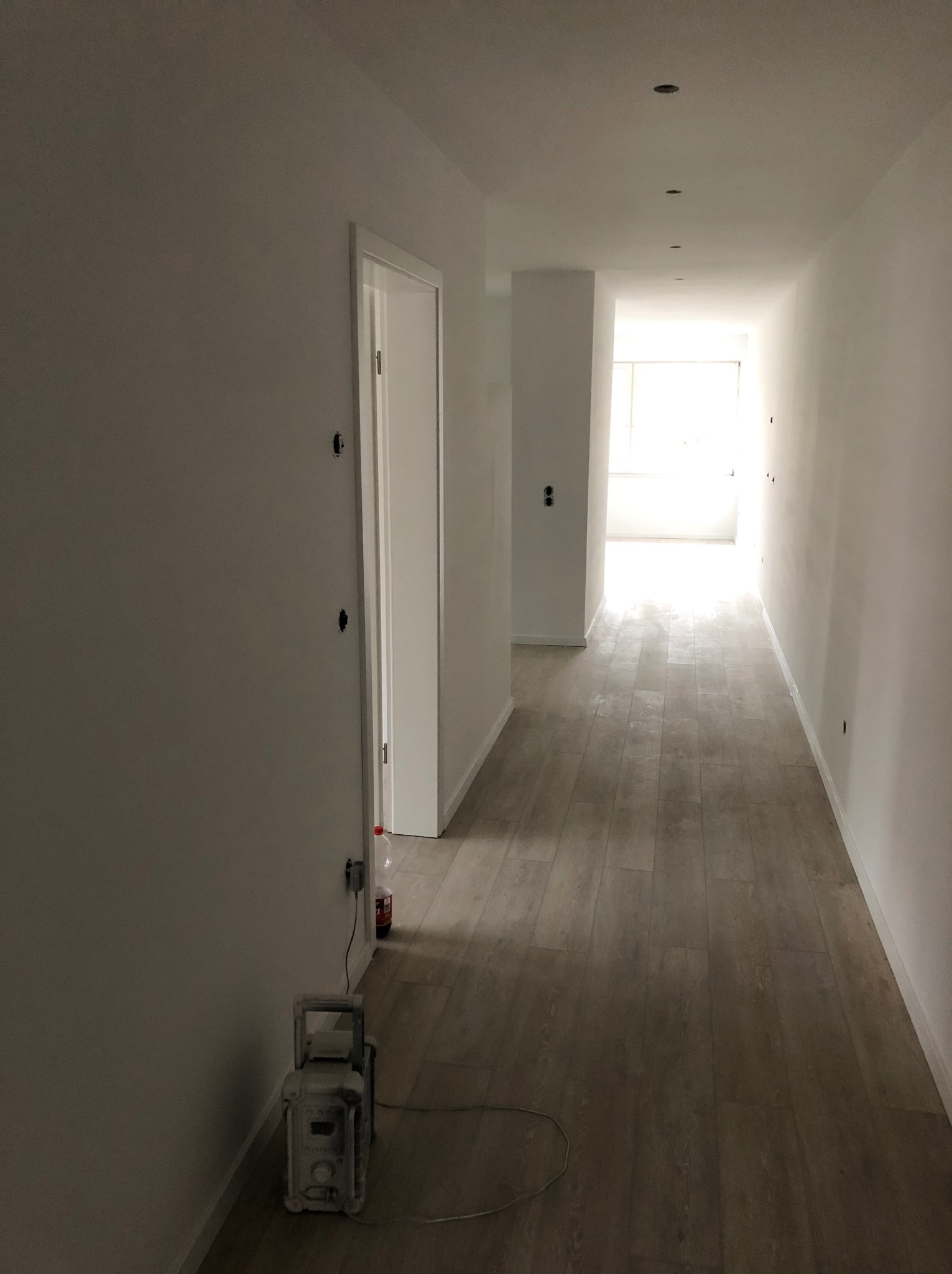 Korytarz w trakcie odświeżania, ściany pomalowane na biało, widoczne ślady po gniazdkach, podłoga wyłożona panelami imitującymi drewno, w rogu pomieszczenia stoi urządzenie do szpachlowania.