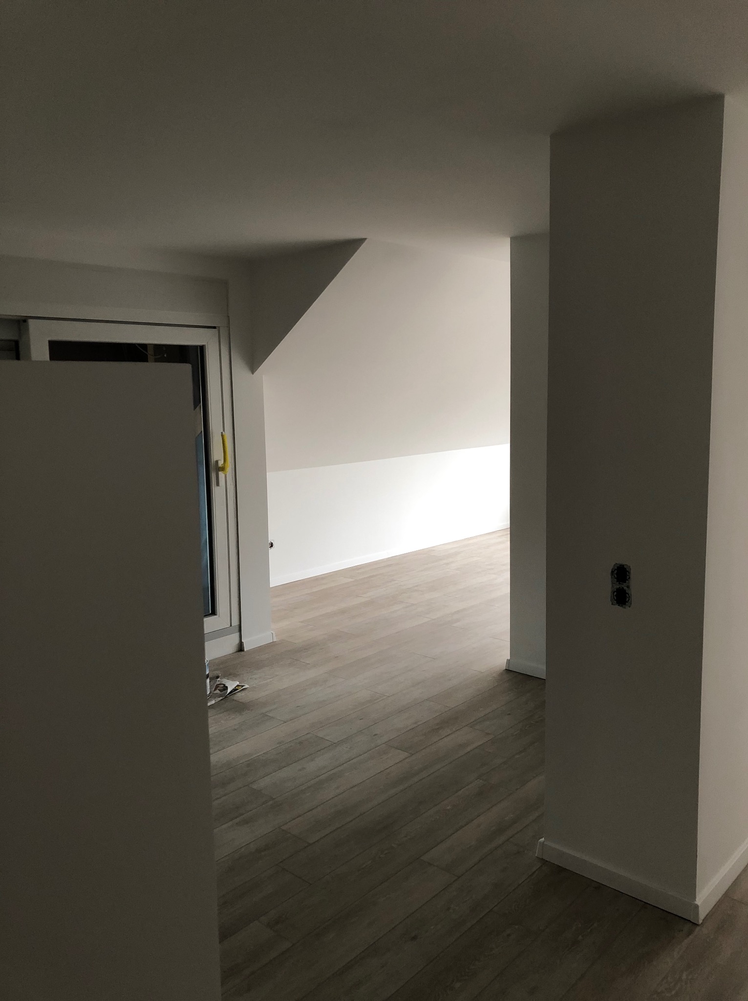 Wnętrze poddasza po remoncie: świeżo pomalowane ściany na biało, drewnopodobna podłoga, widoczne gniazdka elektryczne bez osprzętu, okno dachowe.