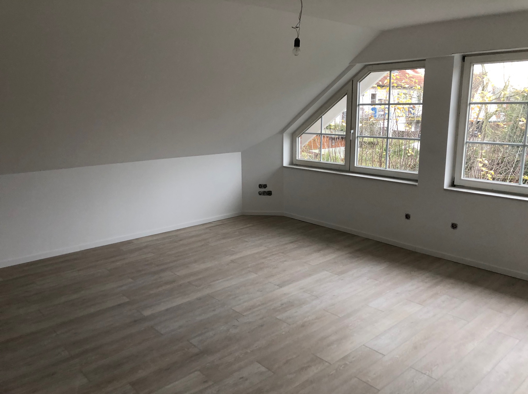 Poddasze po remoncie z jasną podłogą imitującą drewno, świeżo pomalowane ściany na biało, dwa okna z widokiem na jesienne drzewa, widoczne gniazdka elektryczne i wisząca żarówka.