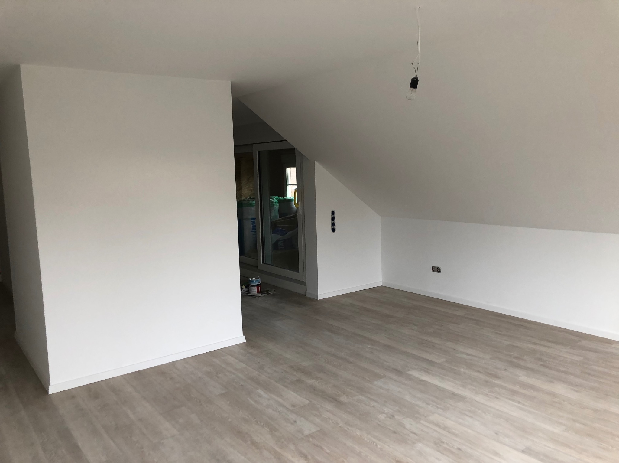 Poddasze po remoncie, ściany pomalowane na biało, panele podłogowe imitujące drewno, widoczne puszki po farbie przy drzwiach balkonowych, goła żarówka zwisająca z sufitu.