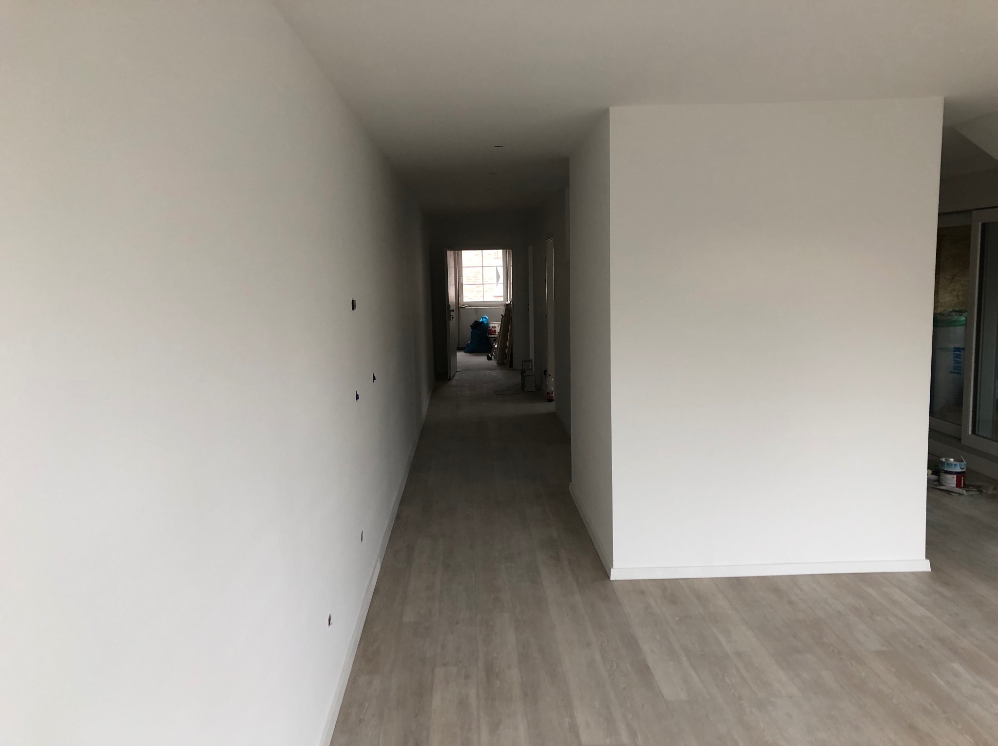 Korytarz w jasnych barwach, świeżo po malowaniu, z widocznymi śladami po otworach na ścianie, podłoga wyłożona panelami imitującymi drewno, w głębi widoczne okno z resztkami materiałów budowlanych.