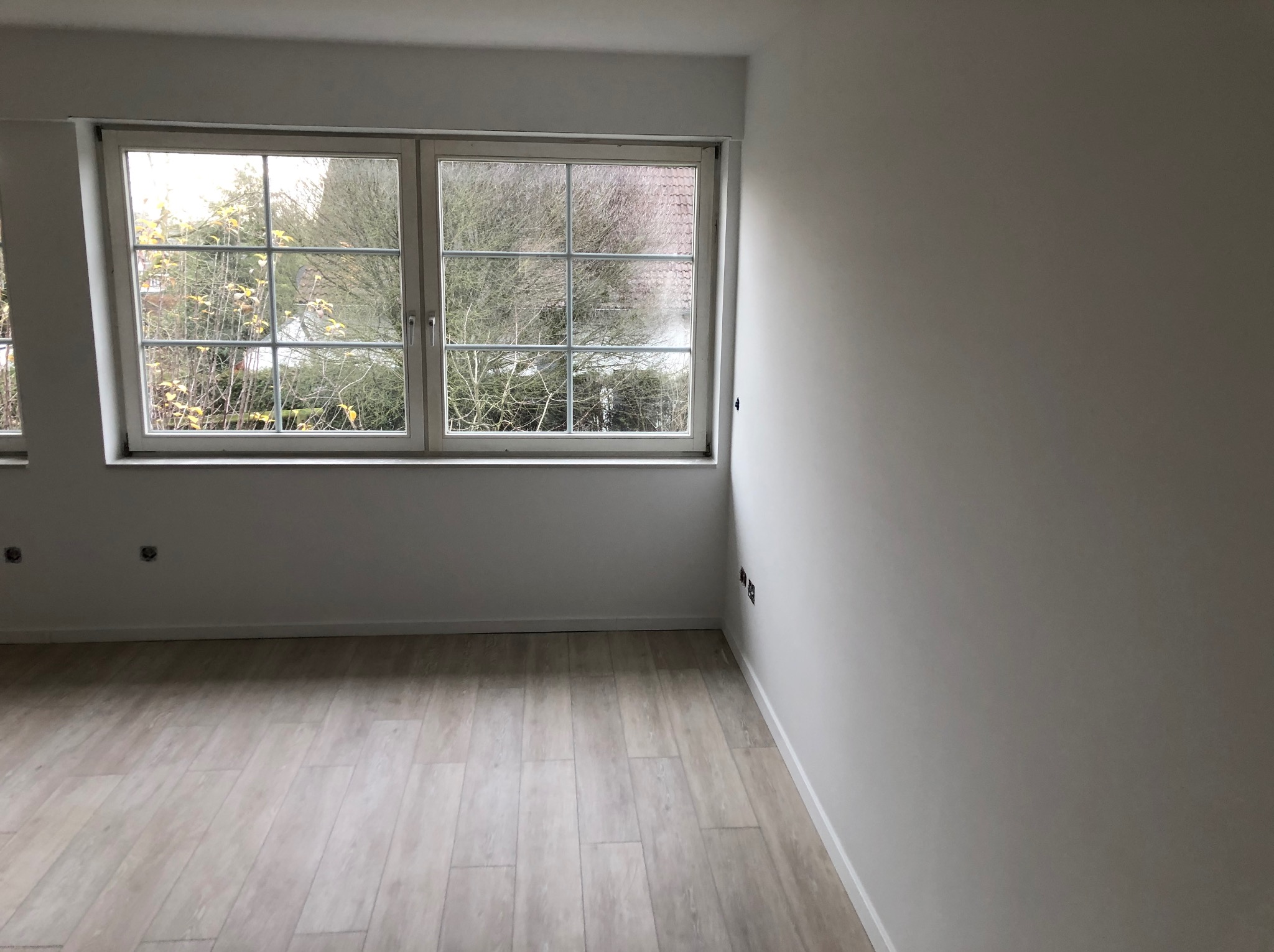 Jasne, nowo wykończone wnętrze pokoju z drewnopodobną podłogą, białymi ścianami i oknem z widokiem na drzewa bez liści.