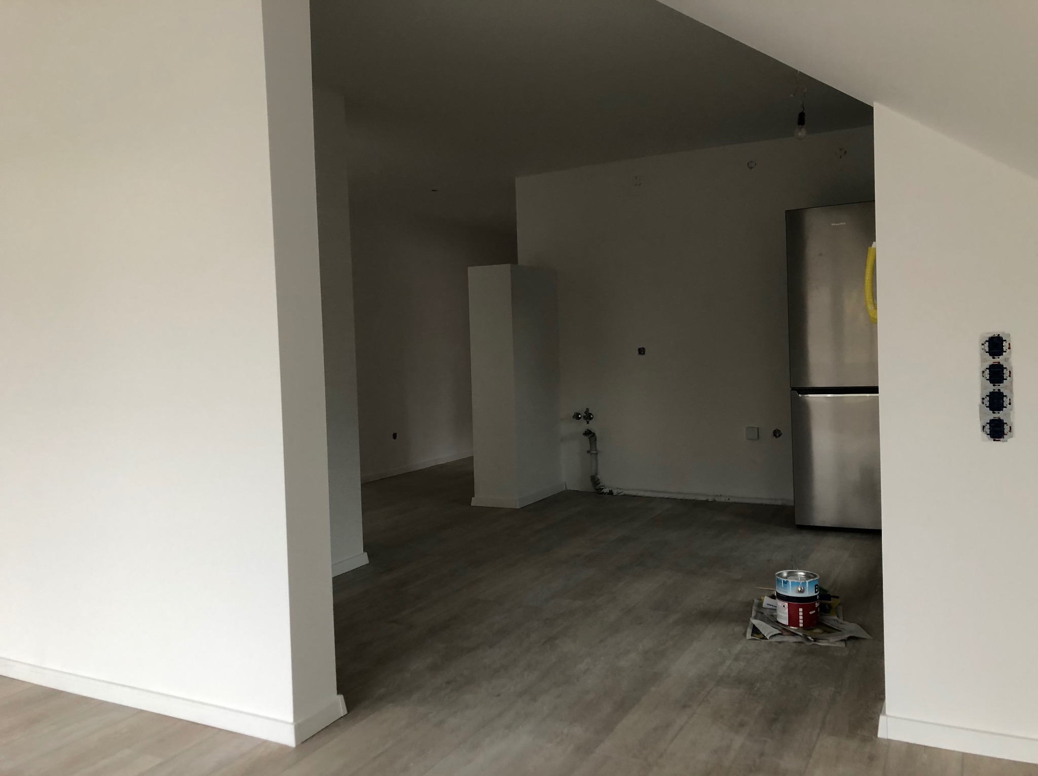 Świeżo pomalowane na biało ściany w przestronnym wnętrzu z drewnopodobną podłogą, widoczne puszki z farbą na podłodze, fragment lodówki i gniazdka elektryczne bez obudowy.