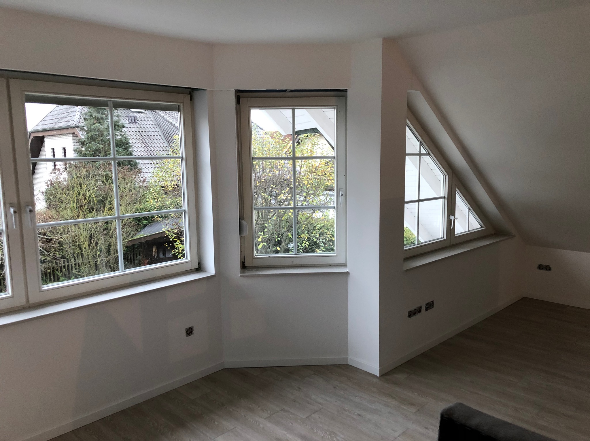 Jasne, wykończone wnętrze poddasza z trzema oknami o różnym kształcie, świeżo pomalowane ściany na biało, panele podłogowe imitujące drewno, widoczne gniazdka elektryczne.