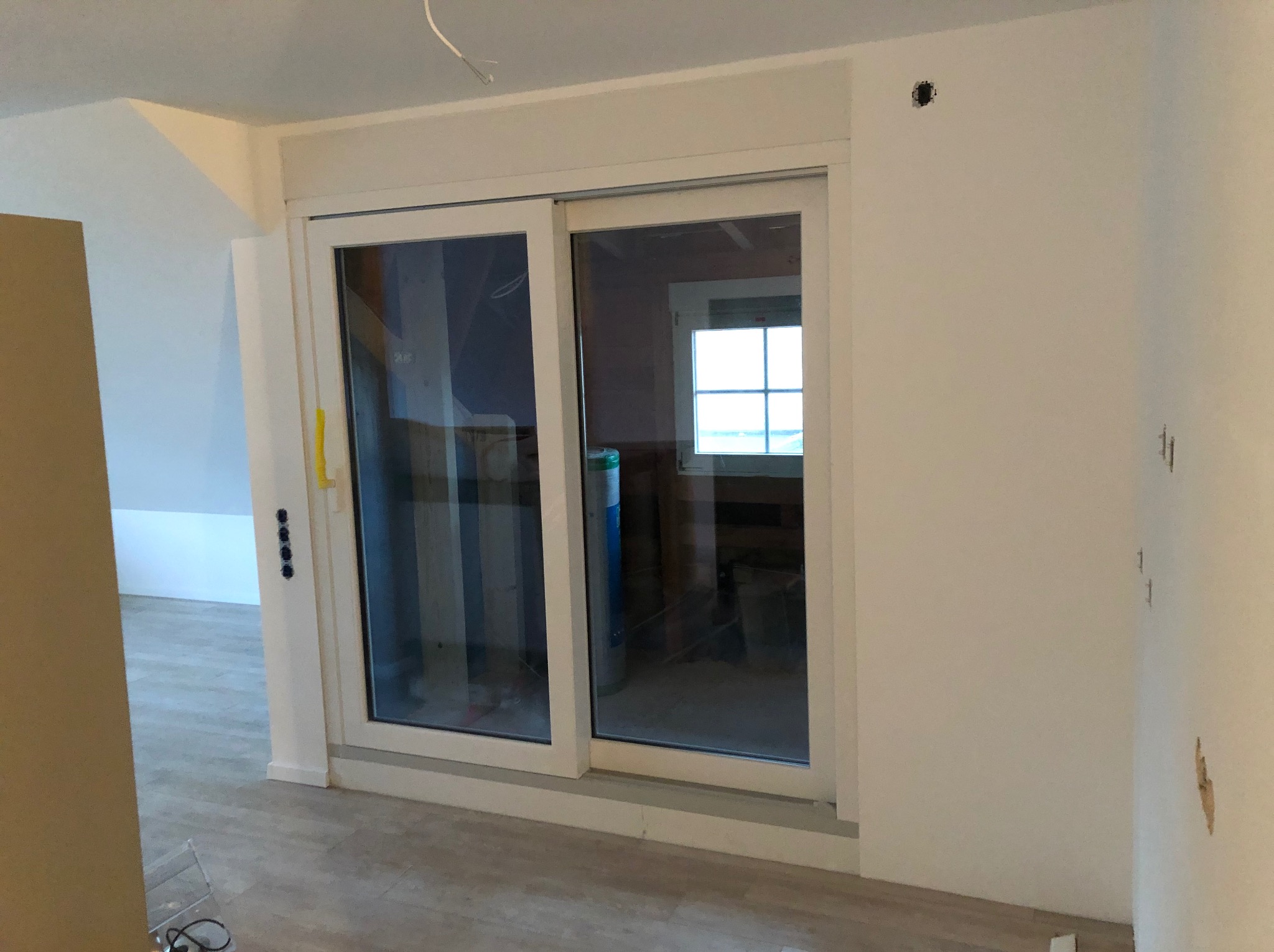 Wnętrze w trakcie prac wykończeniowych z zamontowanymi przesuwnymi drzwiami balkonowymi w białej ramie, widoczne ślady po szpachlowaniu na ścianie i otwór na gniazdko elektryczne.