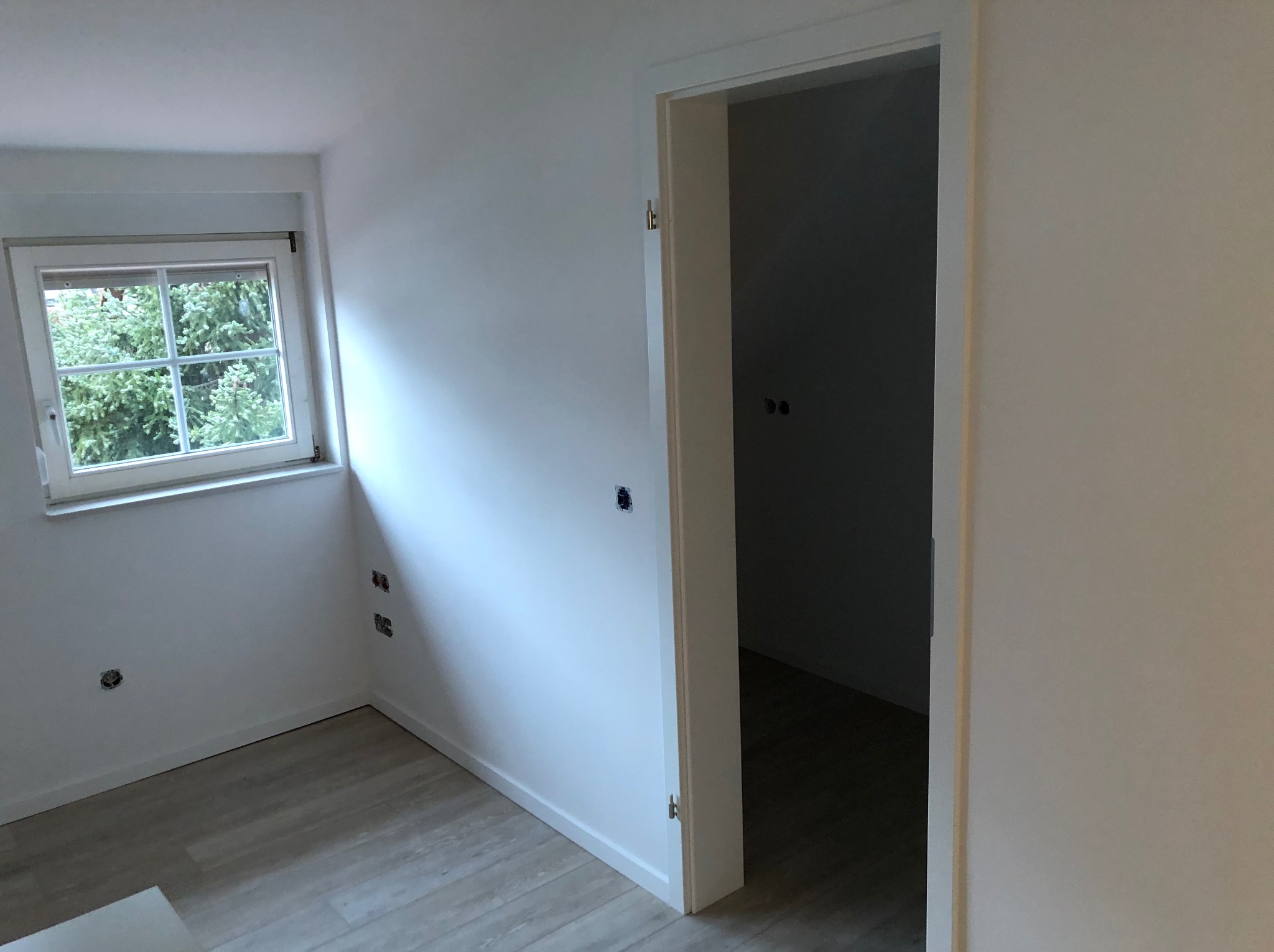 Świeżo pomalowane na biało wnętrze pokoju z drewnopodobną podłogą, widoczne gniazdka elektryczne bez osprzętu i otwarte białe drzwi do ciemniejszego pomieszczenia.