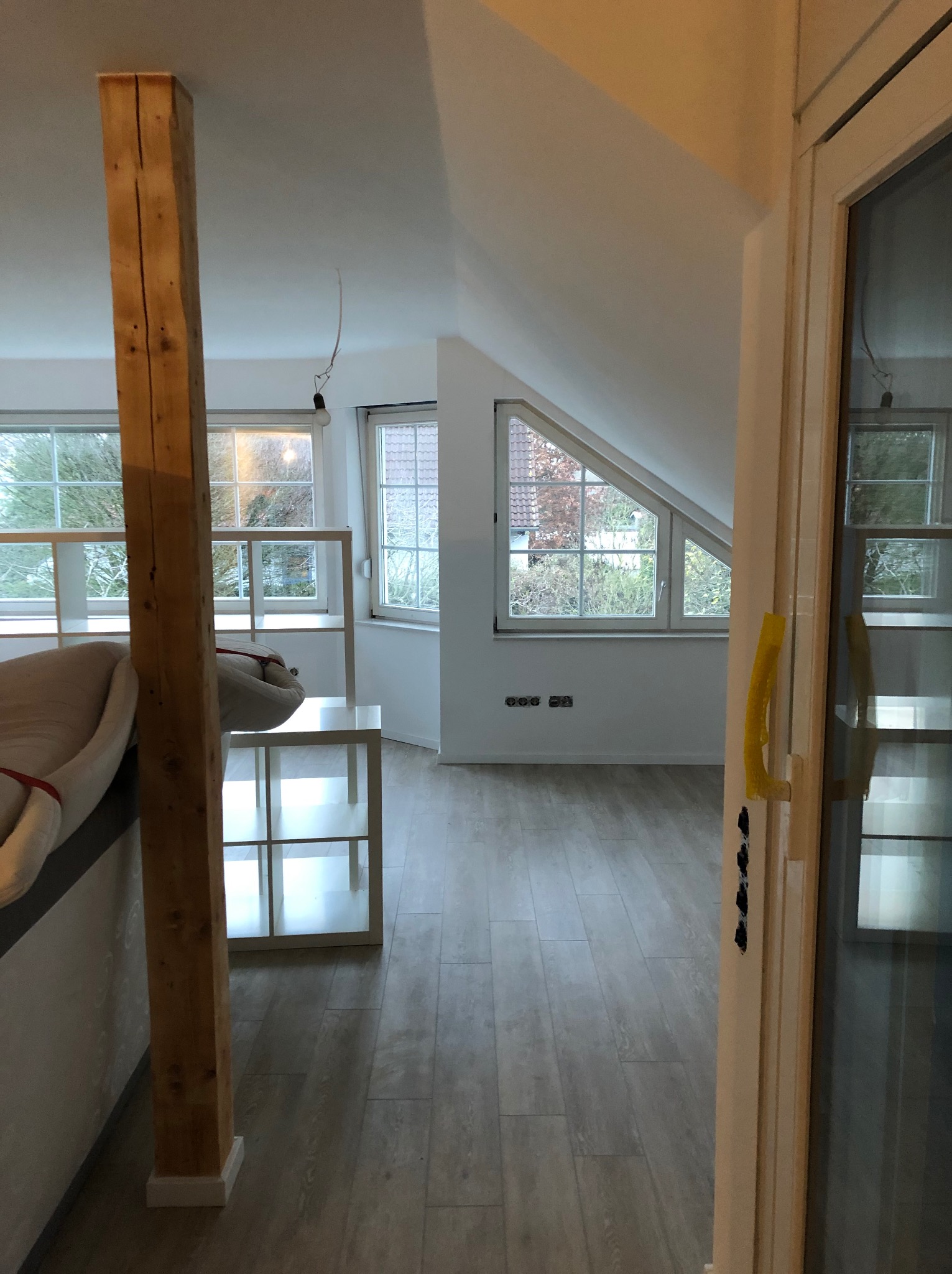 Jasne poddasze z widocznym drewnianym słupem konstrukcyjnym, oknami dachowymi i fragmentem drzwi z żółtą taśmą zabezpieczającą