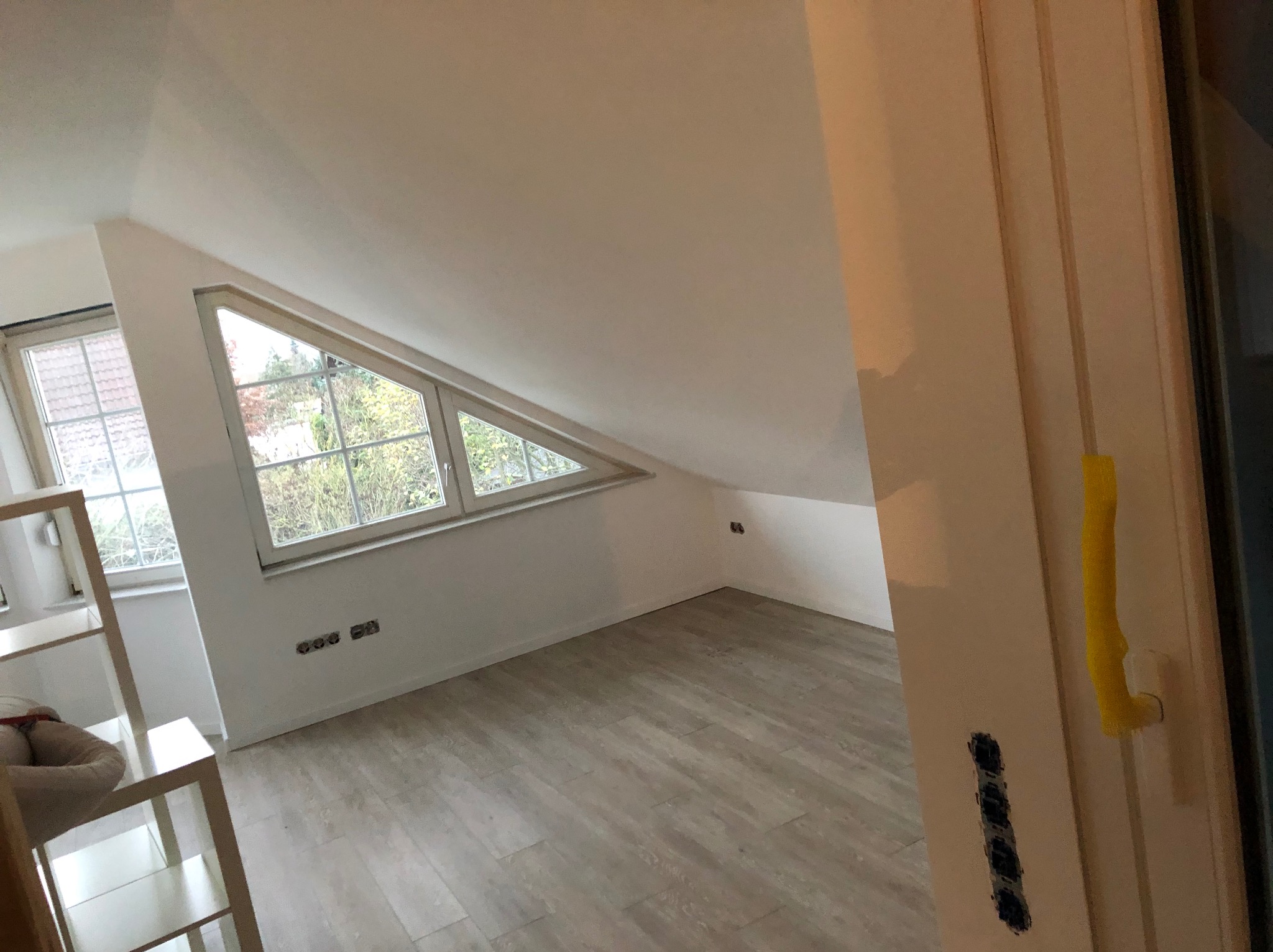 Poddasze po remoncie: białe ściany, panelowa podłoga, trójkątne okno z widokiem na zewnątrz, widoczne gniazdka elektryczne, częściowo widoczny regał z zabawkami i fragment ościeżnicy z widocznymi...