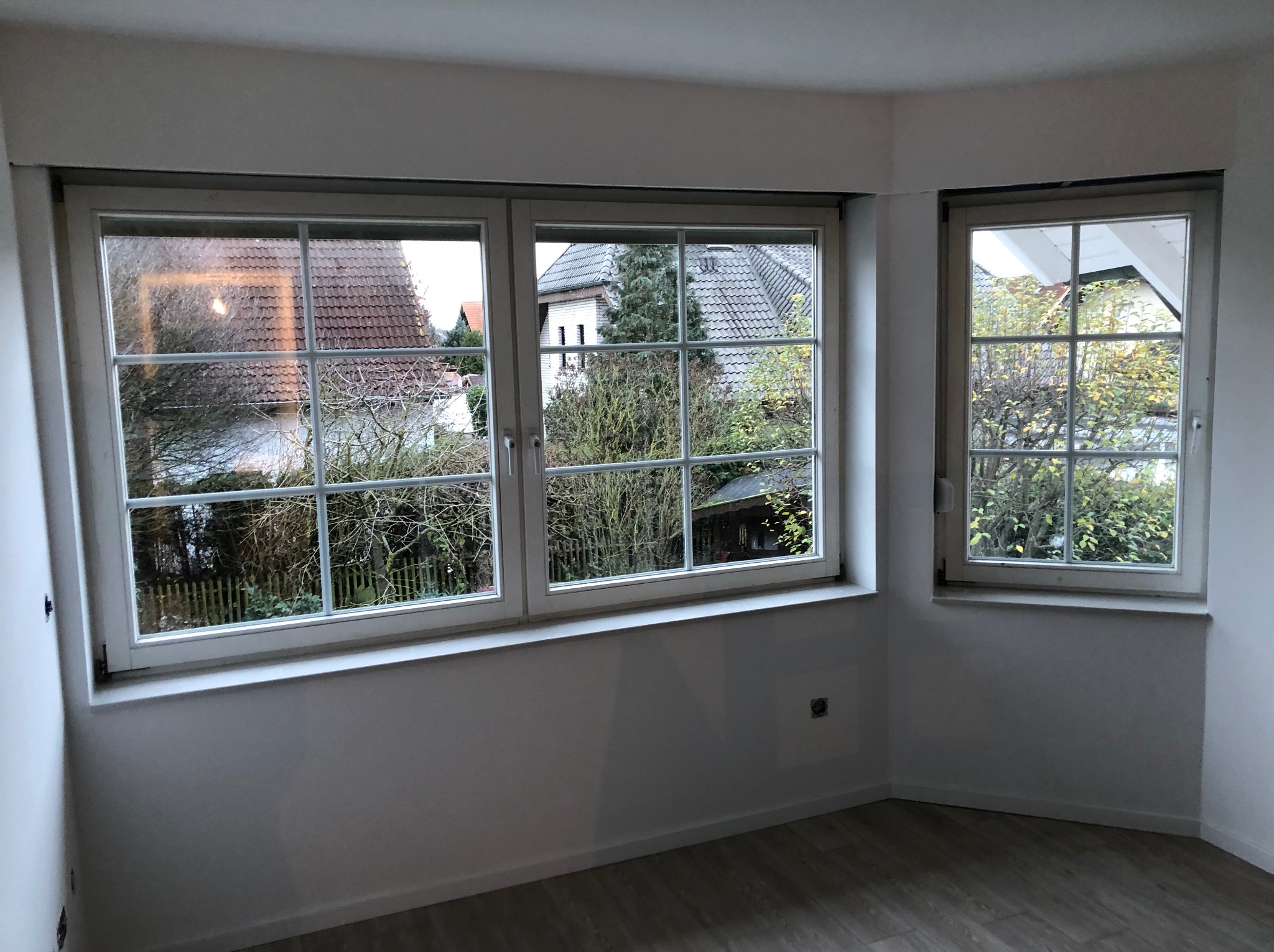 Pusty, świeżo wyremontowany pokój z dwoma oknami z widokiem na domy i drzewa, pomalowany na biało, z panelami podłogowymi o wzorze drewna.