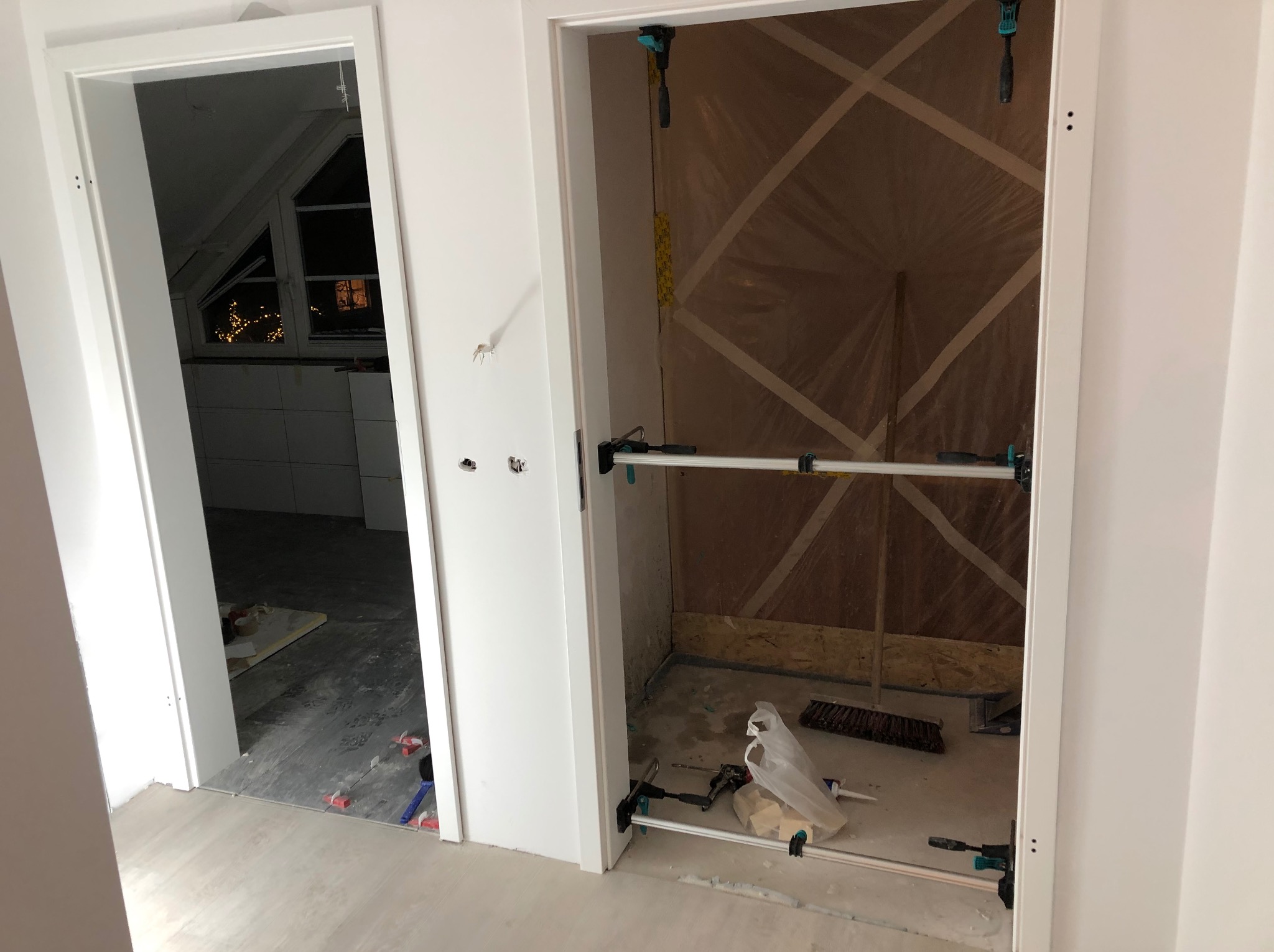 Dwa białe ościeżnice drzwiowe w trakcie montażu, widoczne narzędzia monterskie i folia zabezpieczająca wewnątrz jednego z pomieszczeń, w tle poddasze z oknem.