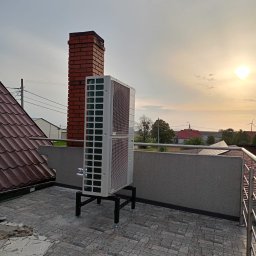 Grupa Majewscy Sp. Z O.O. - Biała pompa ciepła na dachu budynku, z ceglanym kominem w tle i zachodzącym słońcem. Widoczna instalacja na czarnym stelażu, balustrada i fragment dachu z dachówką.