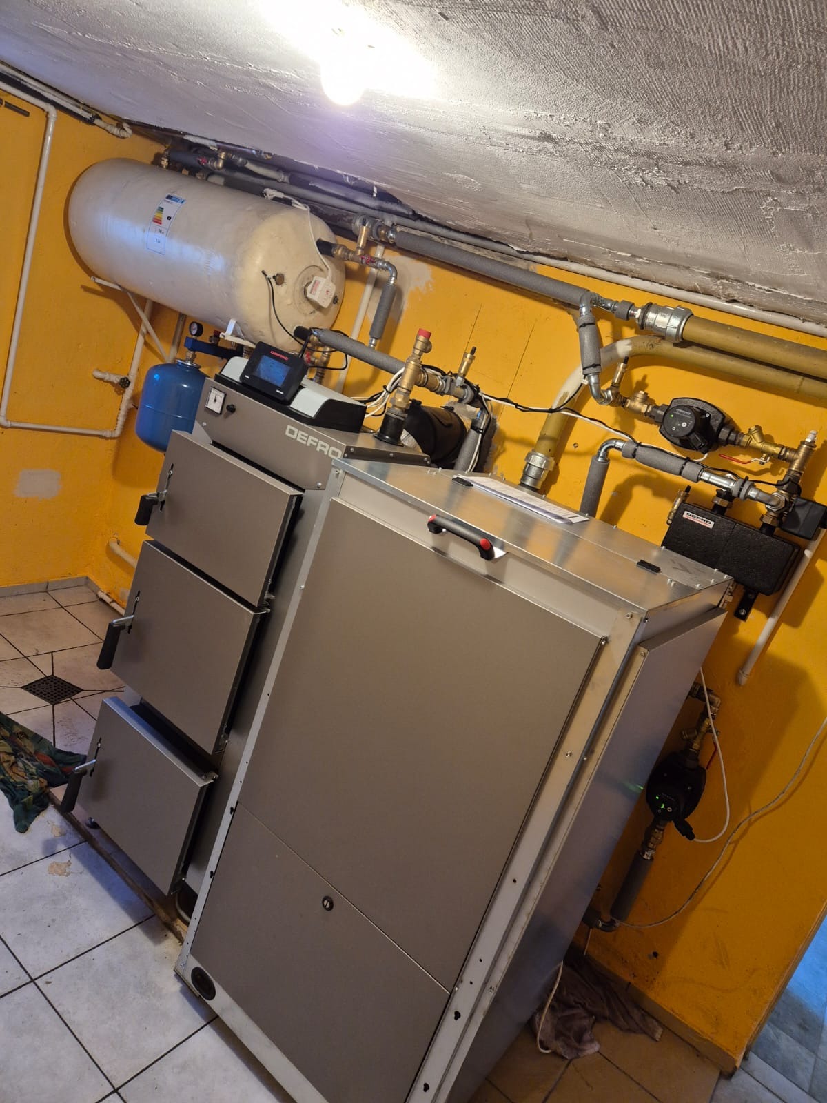 Szary kocioł DEFRO na pellet, z podłączonymi rurami i zbiornikiem, w kotłowni z żółtymi ścianami i białą instalacją. Ujęcie pod kątem.